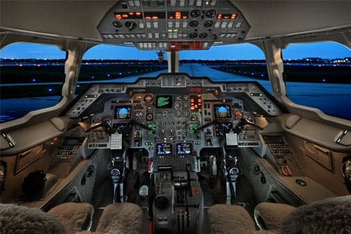 A detailed view of an aircraft cockpit at dusk, looking out onto a runway with lights.