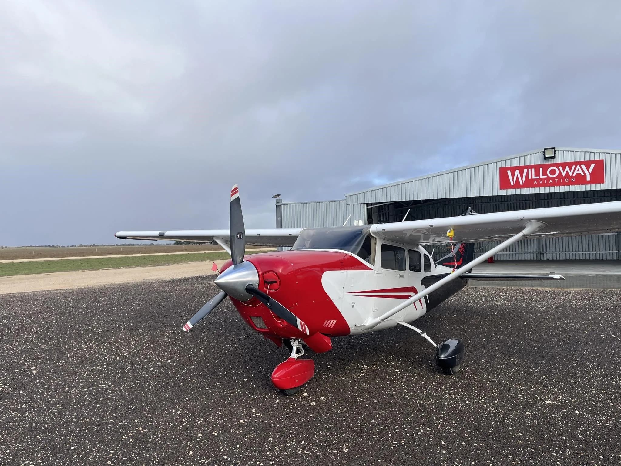 A red and white single-engine propeller aircraft is parked on an airfield in front of a WilloWay Aviation hangar under an overcast sky.