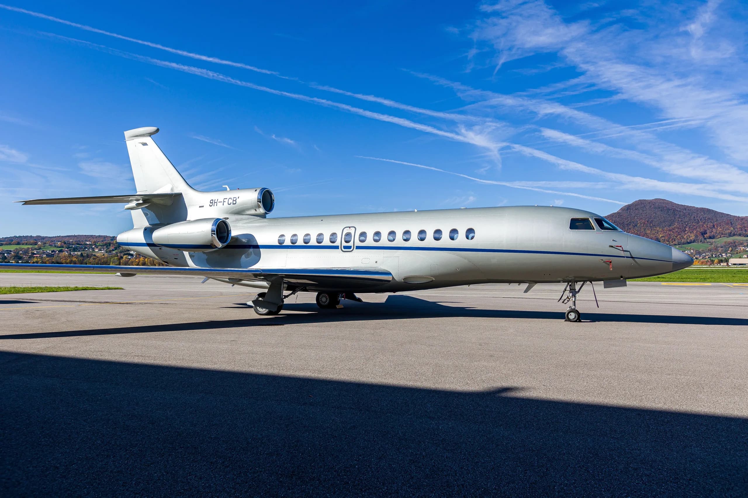 A silver Dassault Falcon 900EX business jet with blue stripes parked on an airfield under a bright blue sky.