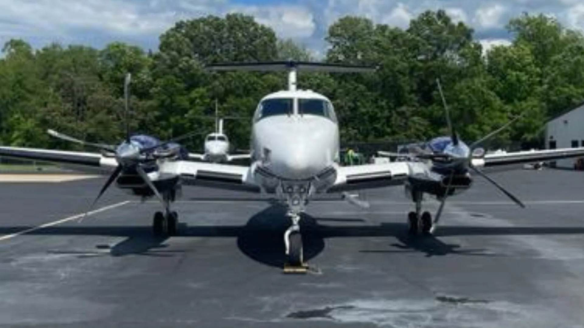 A white and blue Beechcraft King Air N828CJ viewed from the front, parked on an asphalt tarmac with trees in the background.