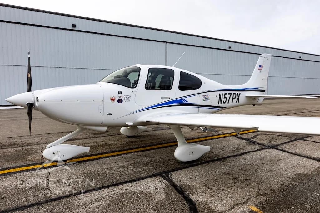 A front-side view of a white Cirrus SR22 aircraft with blue and black stripes, tail number N57PX, parked on an airport tarmac.