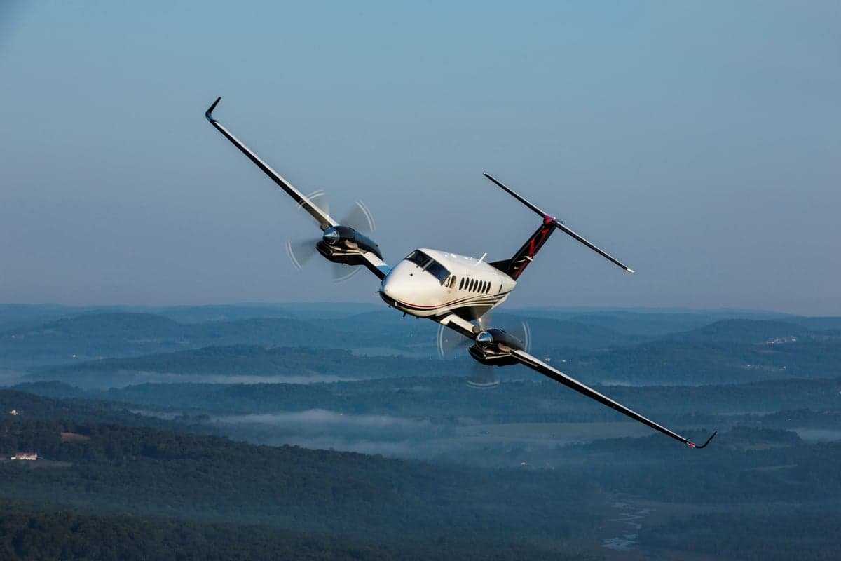 A white and black King Air 350i banks left over a mountainous landscape with a clear sky.