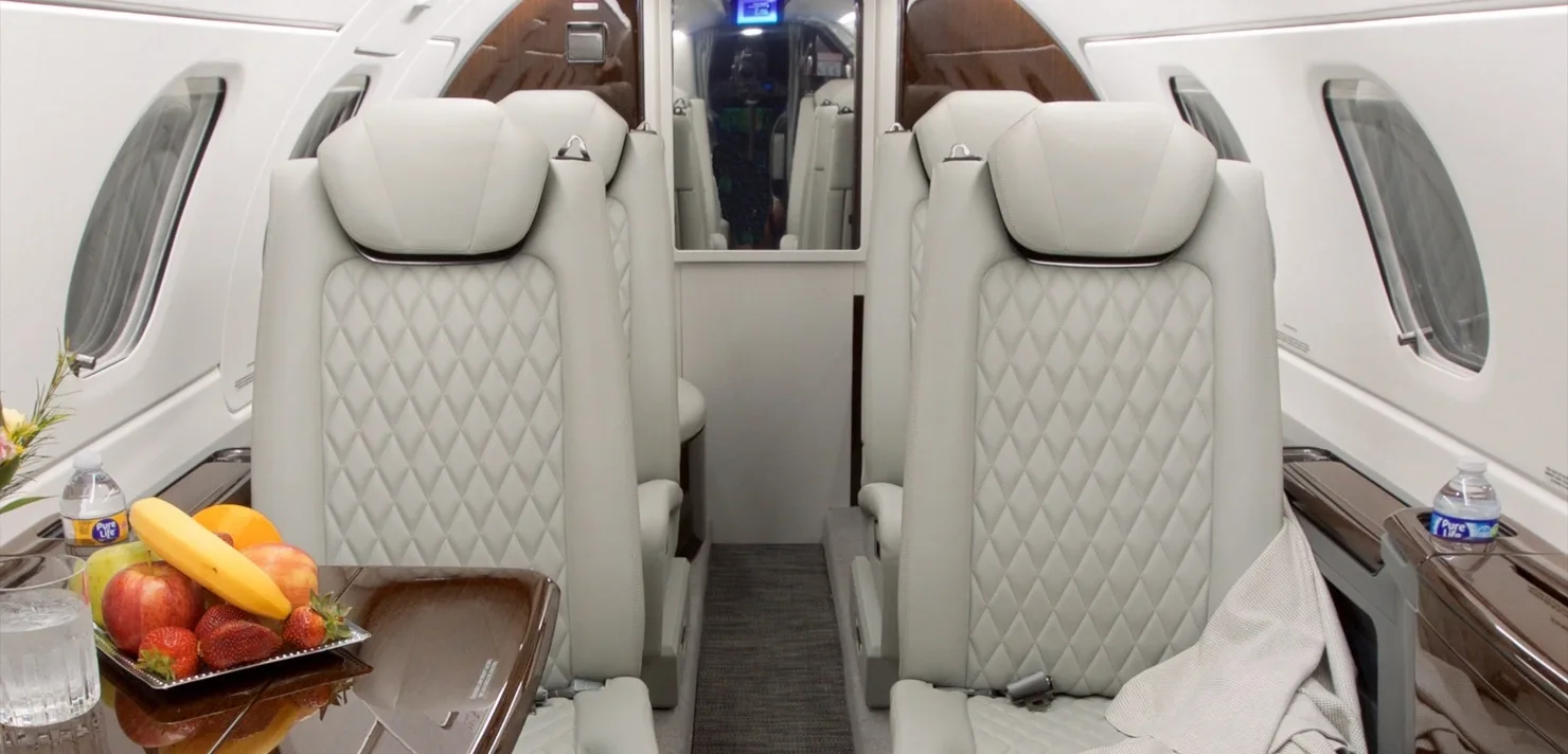 The luxurious cabin interior of the Embraer Phenom 300E, featuring light-colored quilted leather seats and a table with fruit.