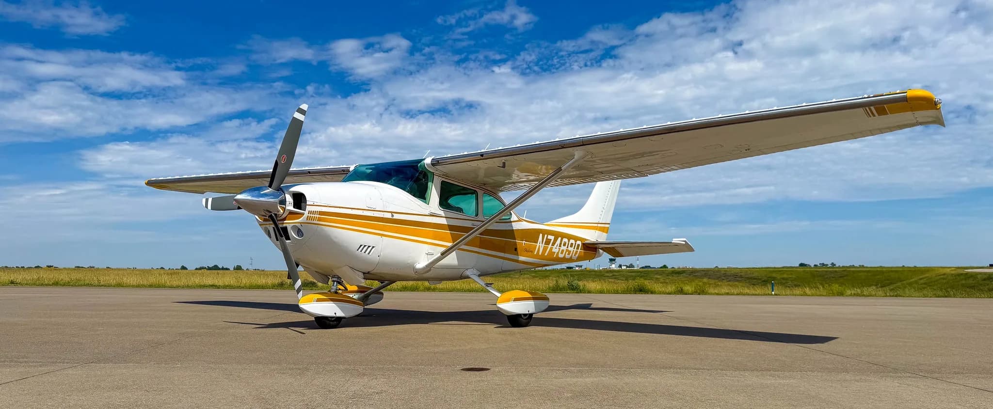 A white and gold Cessna Skylane N7489Q is parked on an airfield tarmac under a partly cloudy sky, viewed from the front-left.