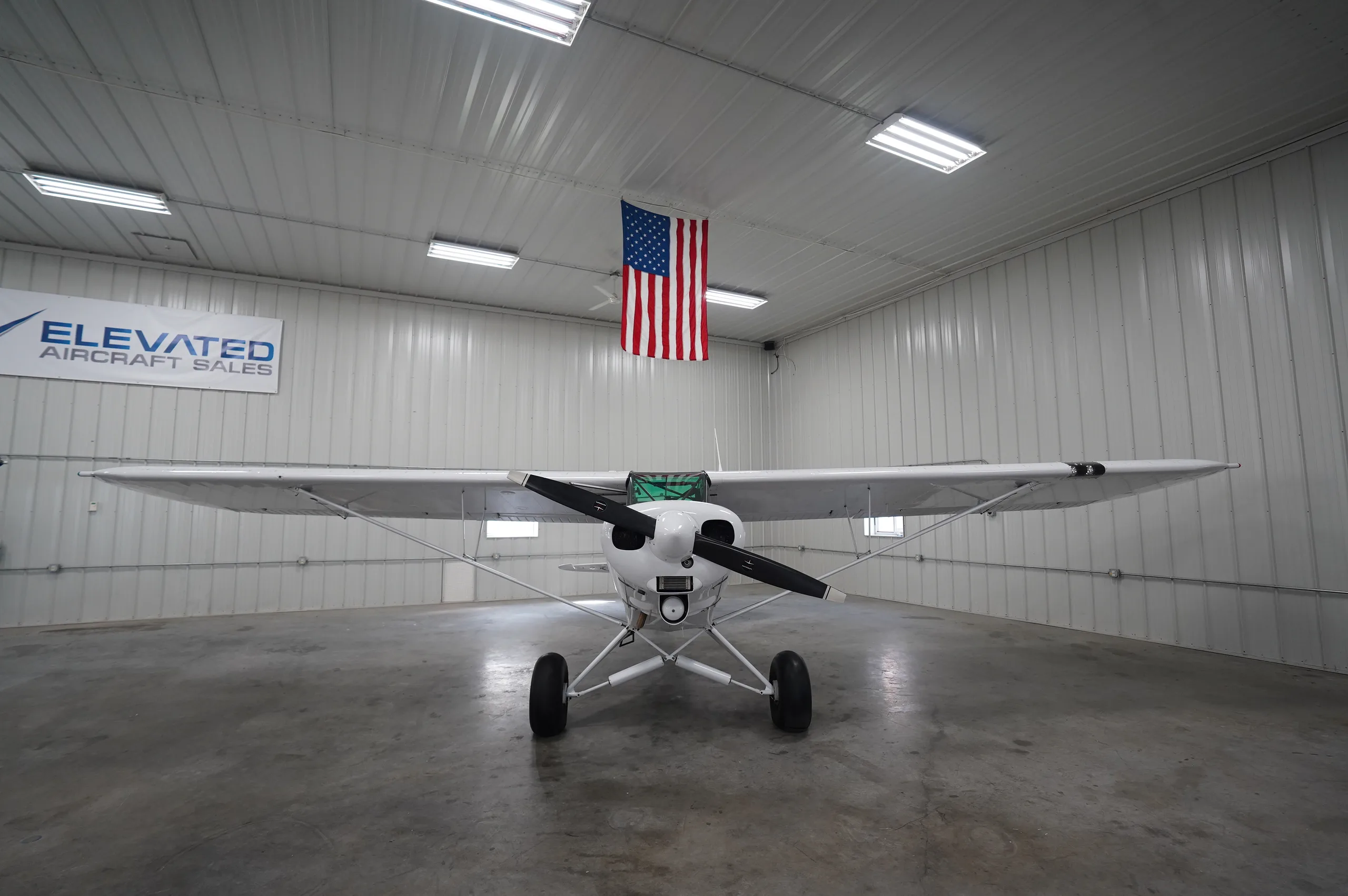 A front view of a white Piper Cub aircraft, registration N4158Y, with red stripes, parked in a hangar under an American flag and "Elevated Aircraft Sales" banner.
