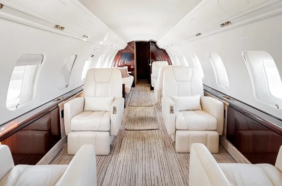 The luxurious aircraft cabin features light cream leather seats arranged in a club configuration with a striped carpet and dark wood accents.