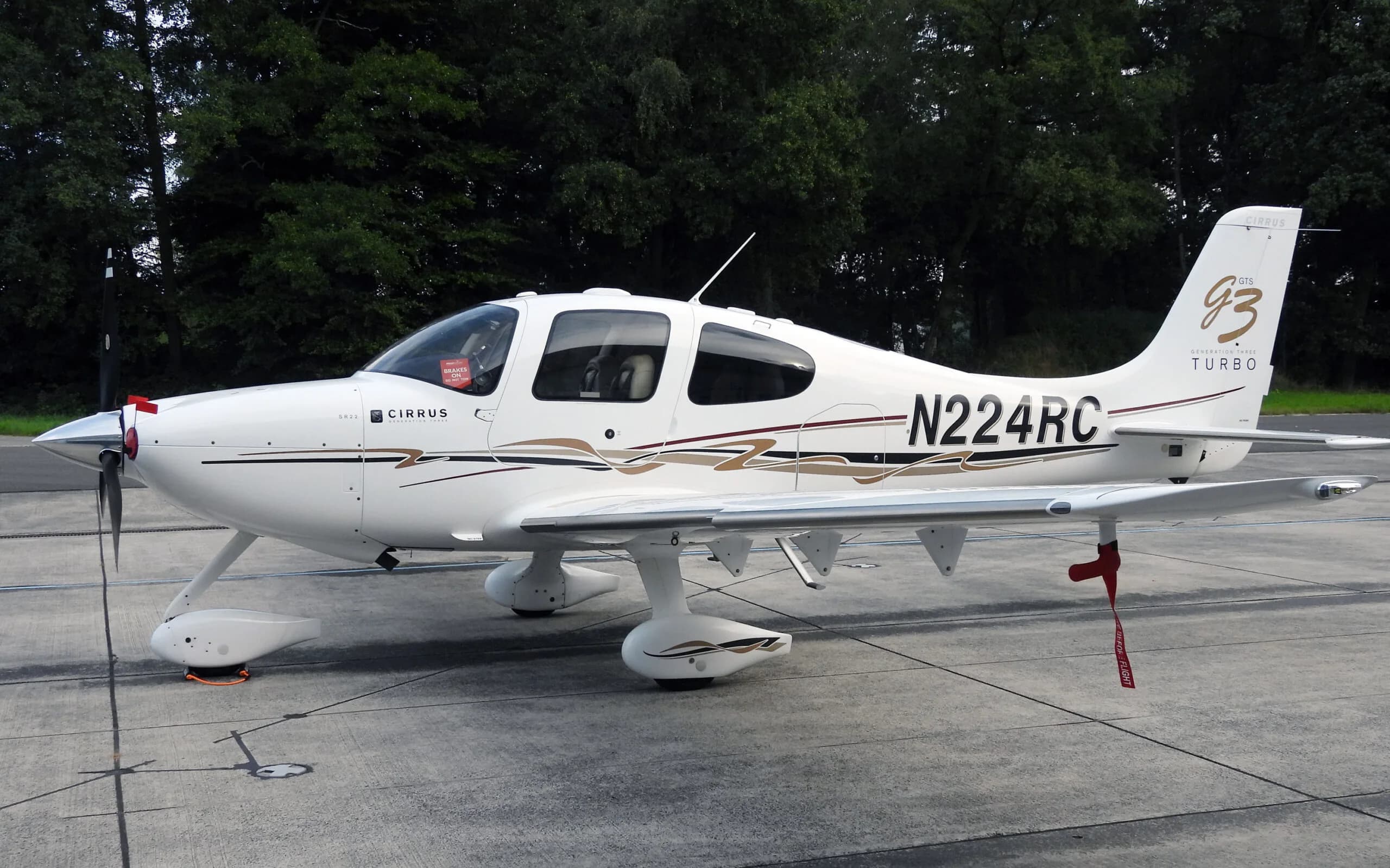 A white Cirrus SR22T (N224RC) with brown and black stripes parked on an asphalt apron with trees in the background.