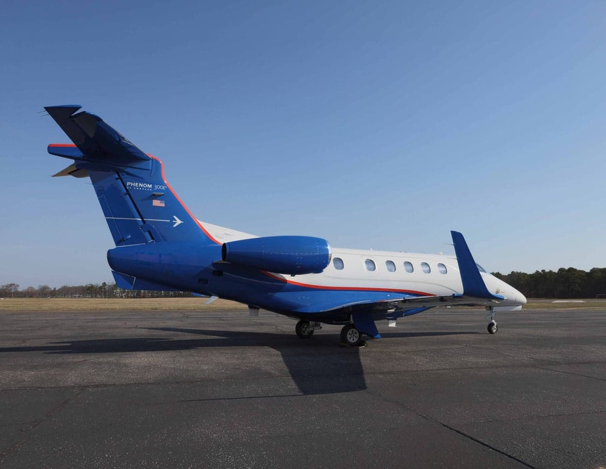 A Phenom 300E aircraft is parked on the tarmac with a blue and white paint scheme.