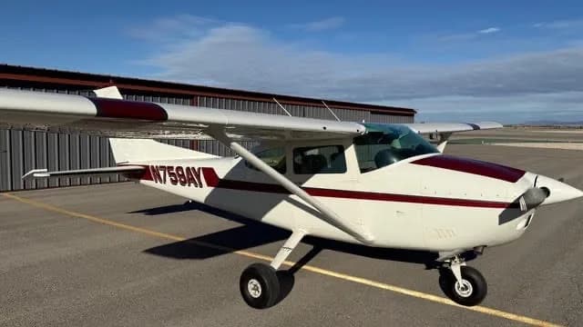 A white single-engine high-wing aircraft with maroon stripes, tail number N759AY, parked on an airfield ramp with a hangar in the background.