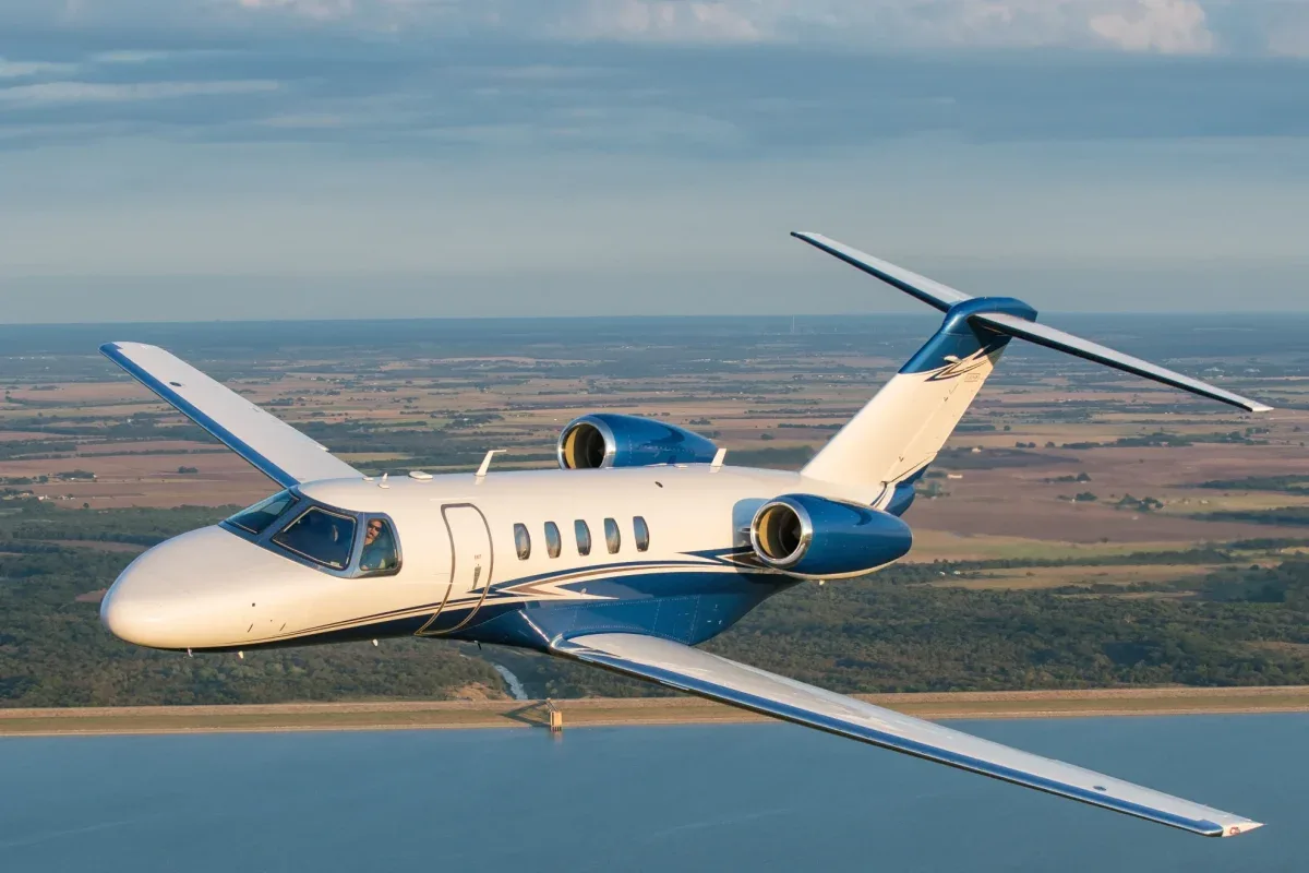 A blue and white Cessna Citation CJ4 flying over a rural landscape.