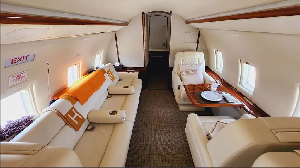 A long view of the aircraft's elegant cabin shows cream leather seating, a sofa with an orange blanket, and a dining table.