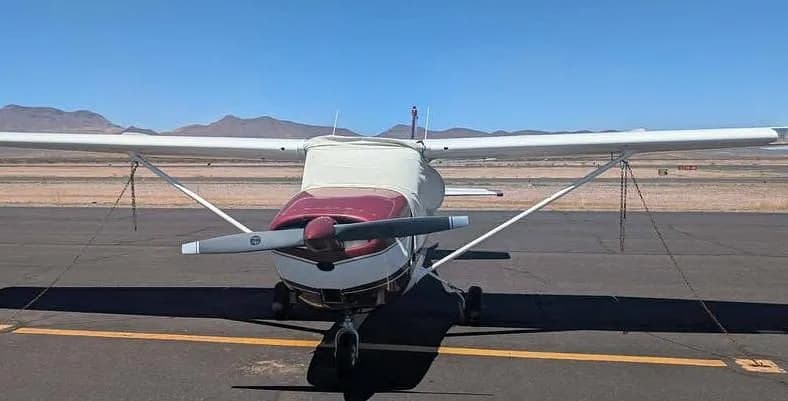 A front view of the white and maroon Cessna 172M, N8452X, on an airfield with a cover over its front section.