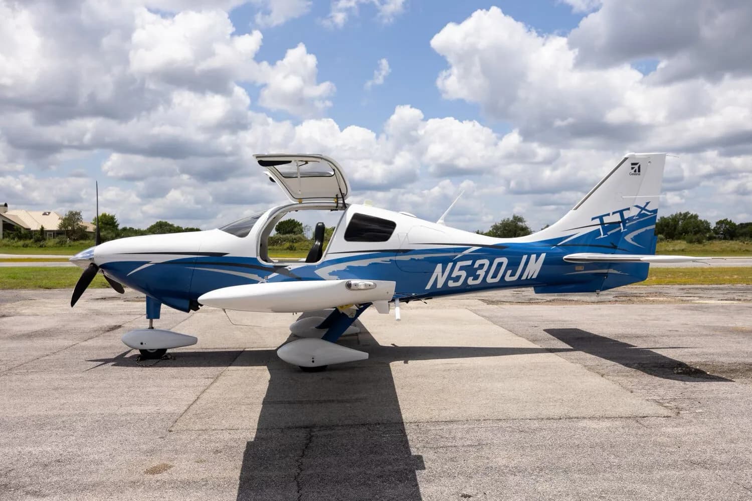 A side view of the white and blue Cessna TTX N530JM with its gull-wing doors open, parked on a tarmac.