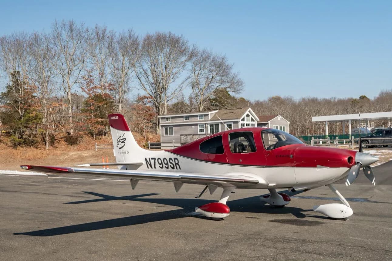 A red and white Cirrus SR22T aircraft, N799SR, parked on an tarmac with trees and a house in the background.