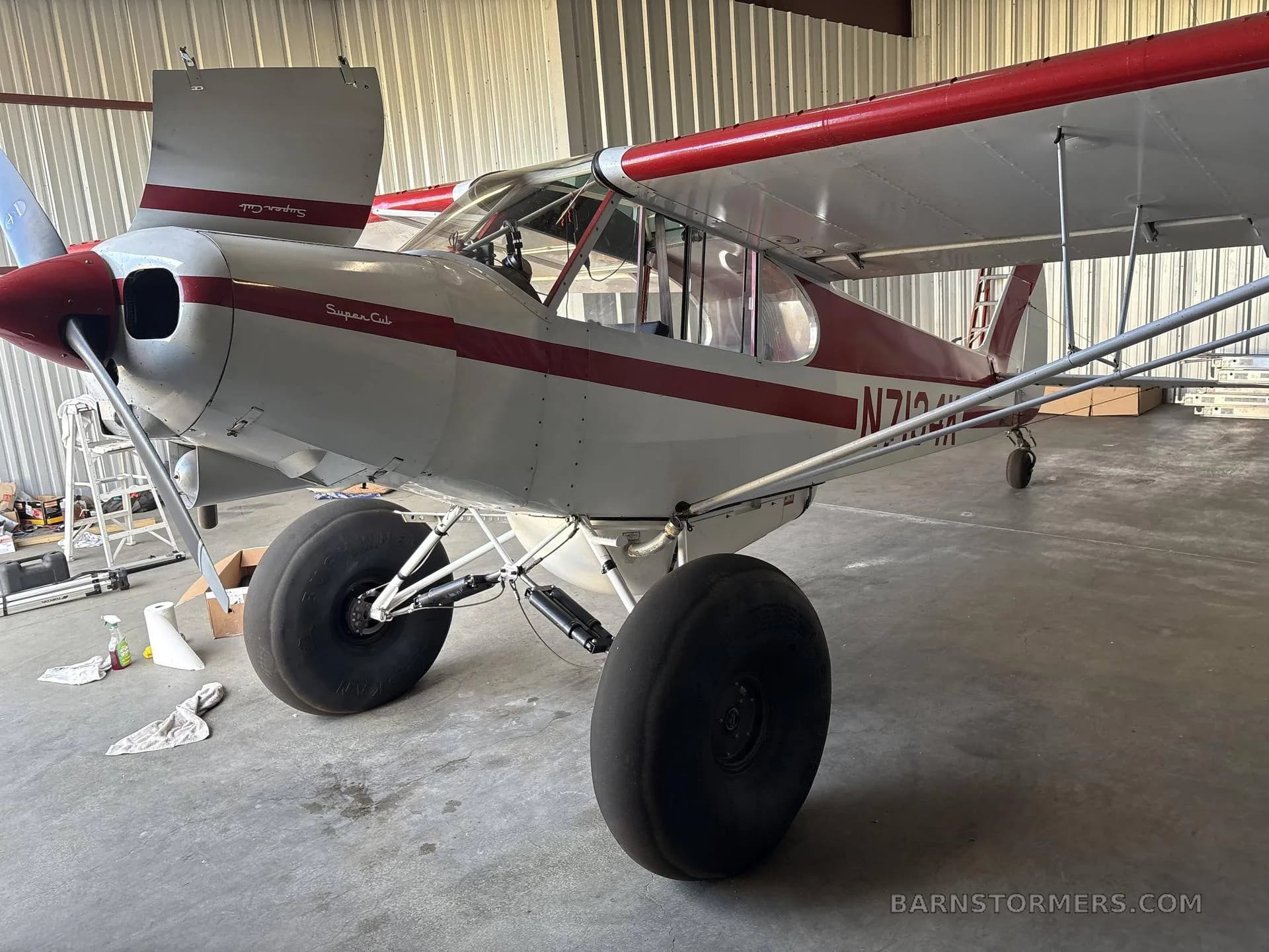 A white and red Piper Super Cub (N7134K) with large tundra tires, parked in a hangar, viewed from the front-right side.