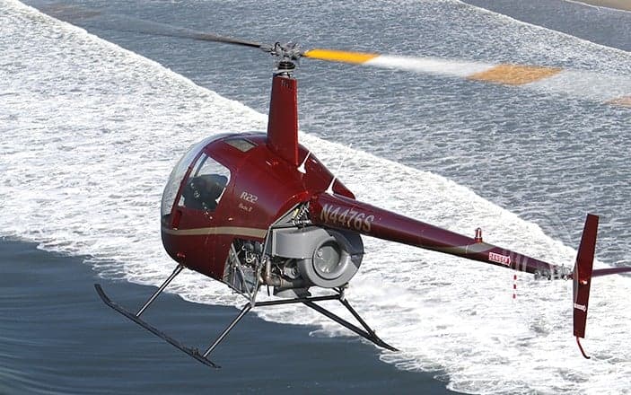 A red Robinson R22 helicopter flying over the ocean.