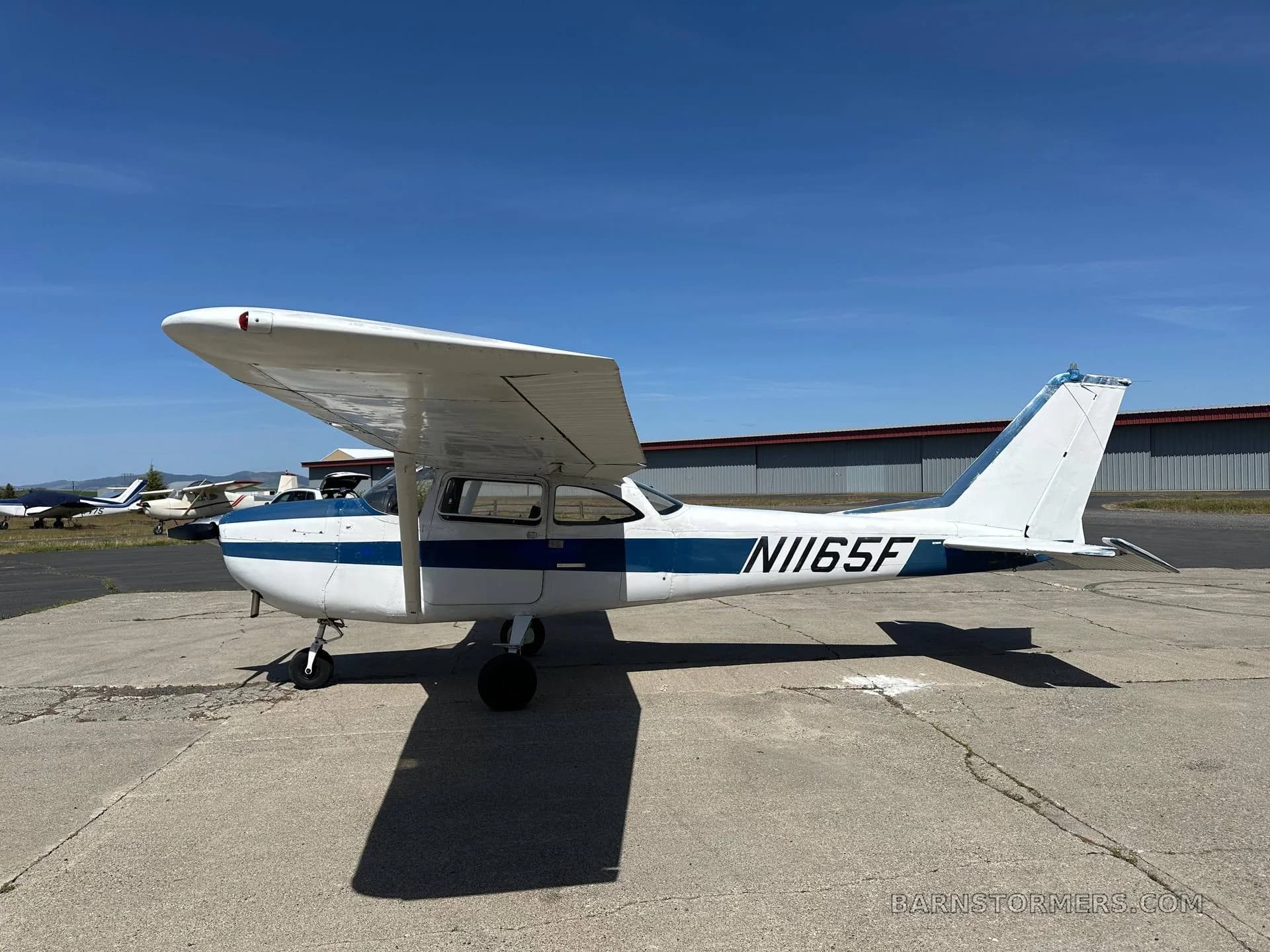 A white and blue Cessna N1165F aircraft is parked on a concrete apron with hangars in the background.