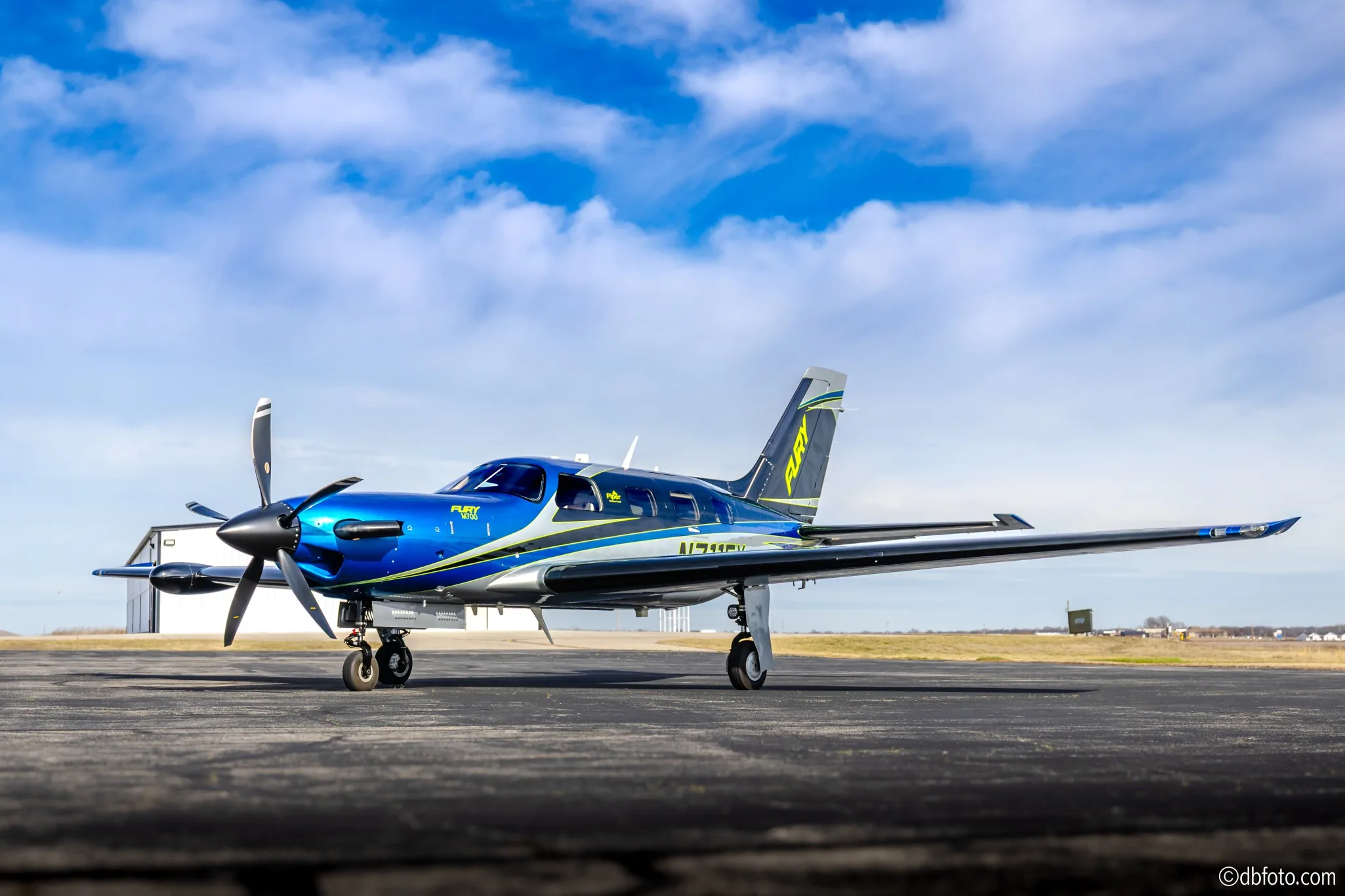 A blue and silver Piper M700 Fury turboprop aircraft (N711FY) parked on a tarmac under a blue sky.