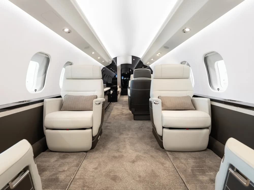 The luxurious interior of a private jet cabin with light grey leather seats and a grey carpeted aisle.