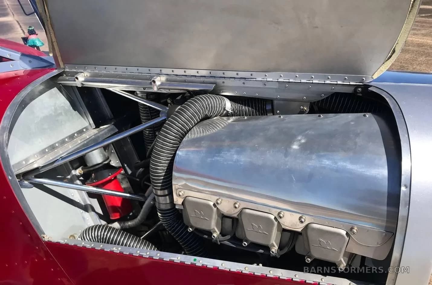 A close-up view of the polished metal engine and components inside the open cowling of a red and silver aircraft.