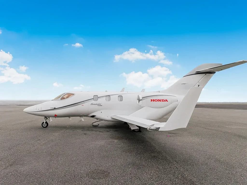 A white HondaJet with grey and red accents, featuring over-wing engines, parked on an asphalt tarmac under a blue sky.