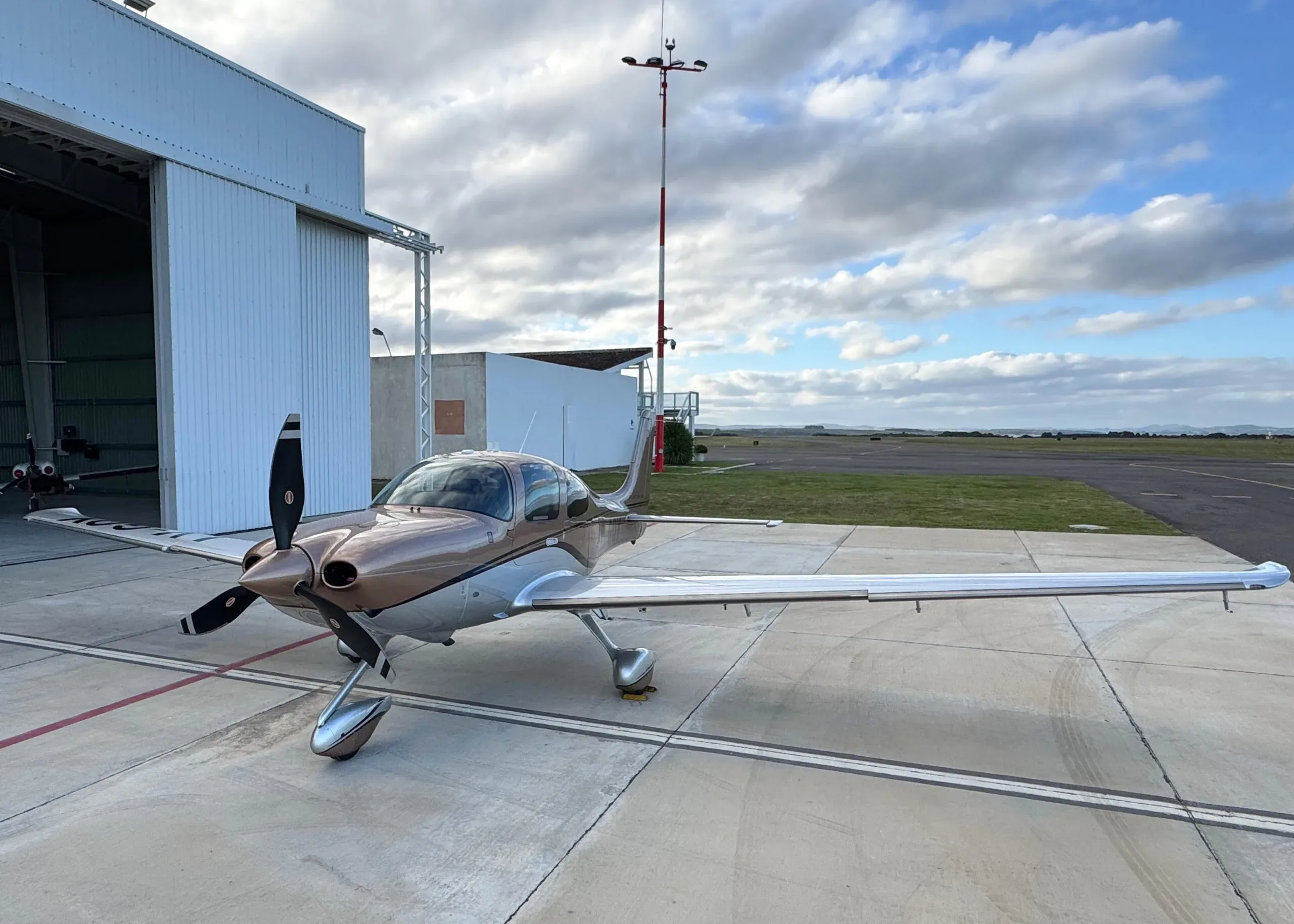 A brown and silver Cirrus SR22T aircraft parked on the tarmac near a hangar with an antenna mast in the background.