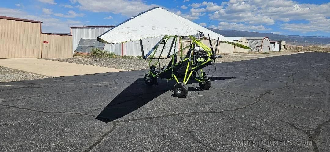 A wide view of a yellow and black ultralight trike with its wing partially folded, parked on an airfield.