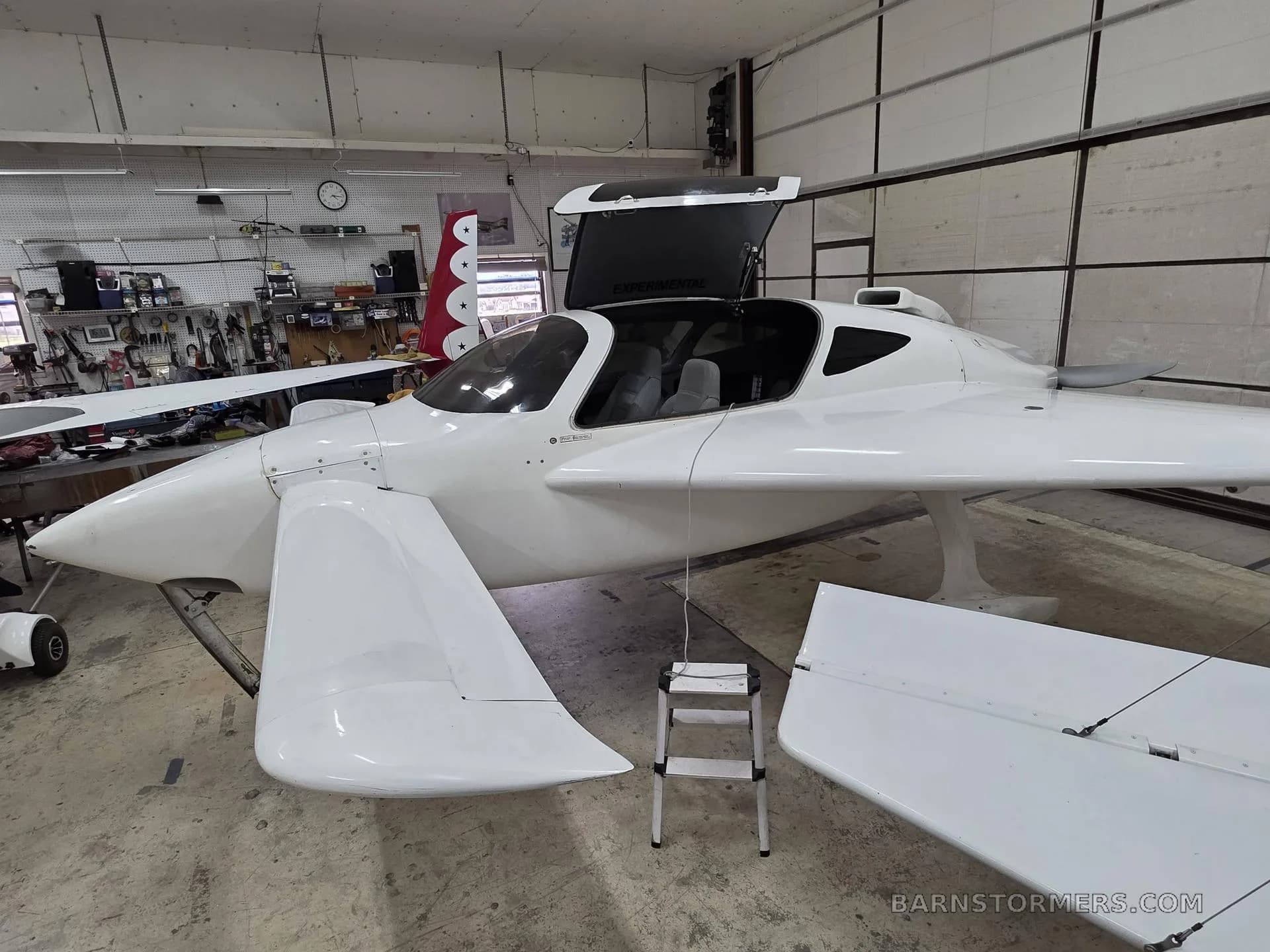 A white experimental aircraft with its canopy open is parked inside a workshop, showing its interior and tools in the background.
