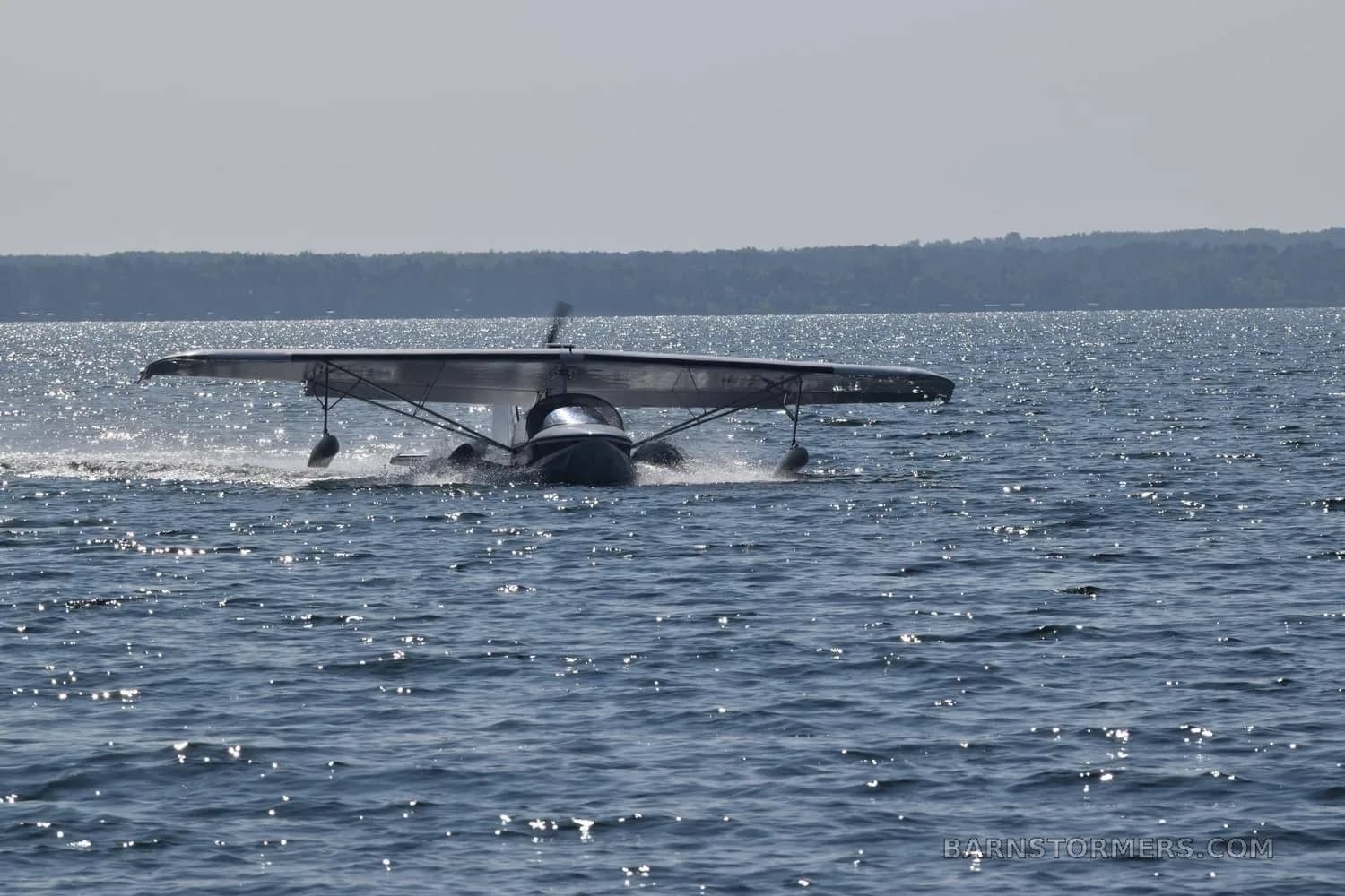 A Searey amphibian aircraft is seen on the water, creating a wake as it moves, with a distant shoreline under a hazy sky.