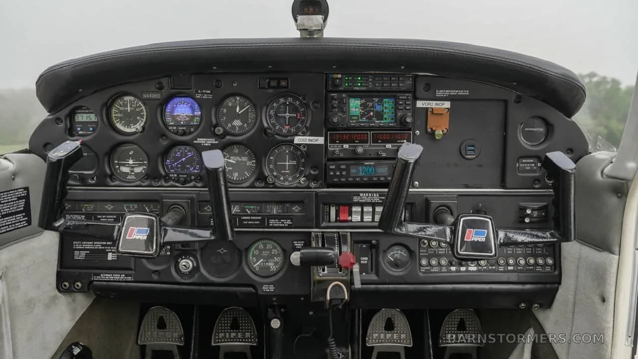 The cockpit of a Piper PA-28 series aircraft, showing the instrument panel with analog gauges and modern avionics, and two control yokes.
