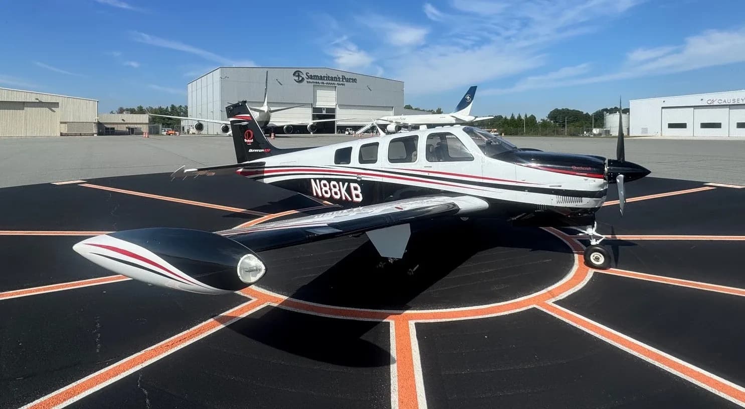 A white and black Beechcraft Bonanza with red stripes, tail number N88KB, parked on an airport tarmac with hangars in the background.