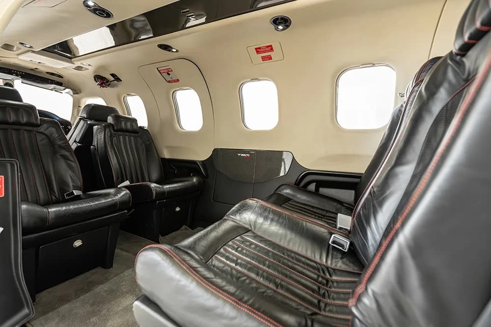 The interior cabin of a Daher TBM 900 aircraft, showing black leather seats with red stitching and large windows.