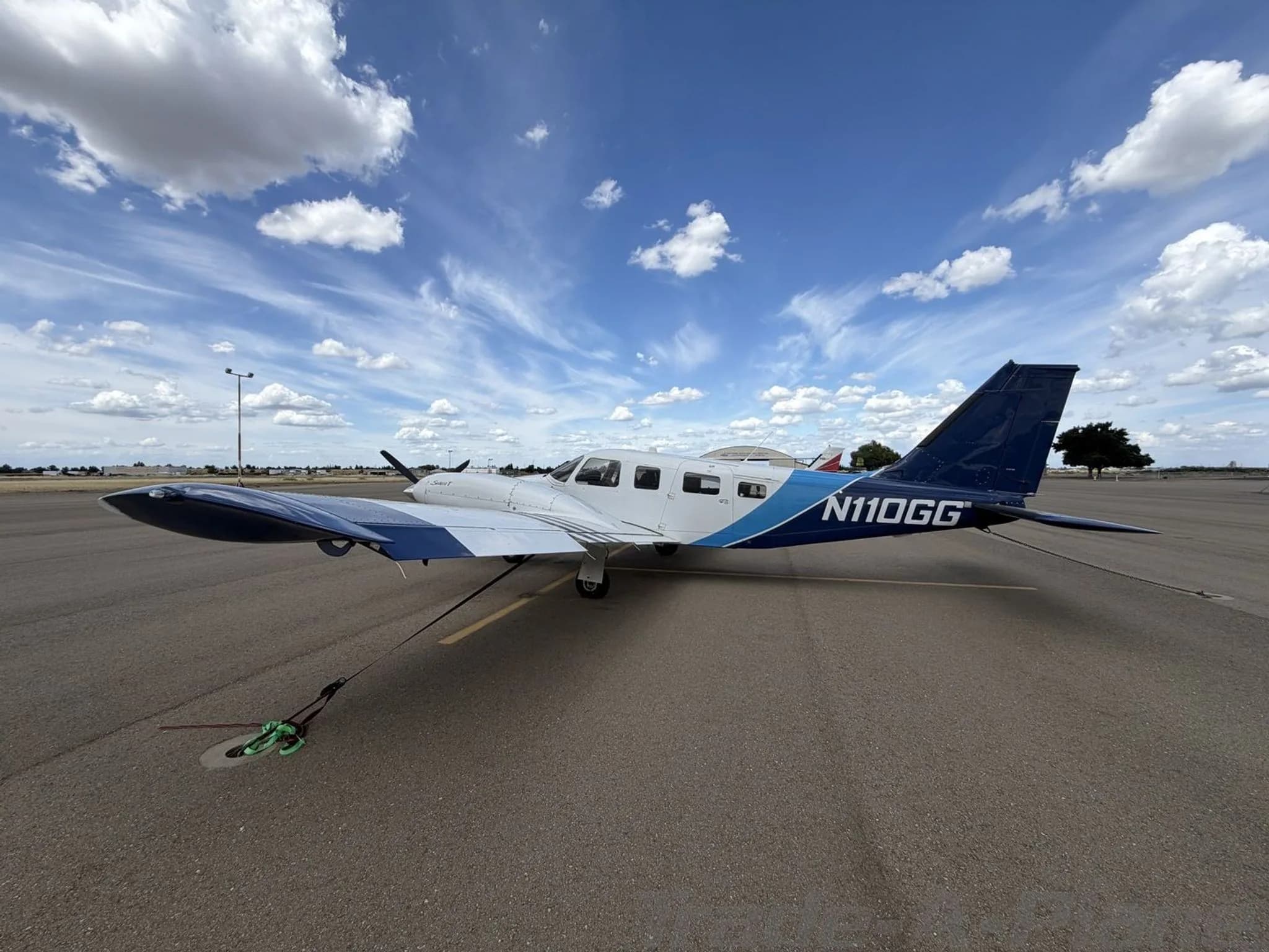 A side-rear view of the white and blue Piper Seneca II twin-engine aircraft, N110GG, tied down on an airport tarmac under a bright blue sky with scattered clouds.