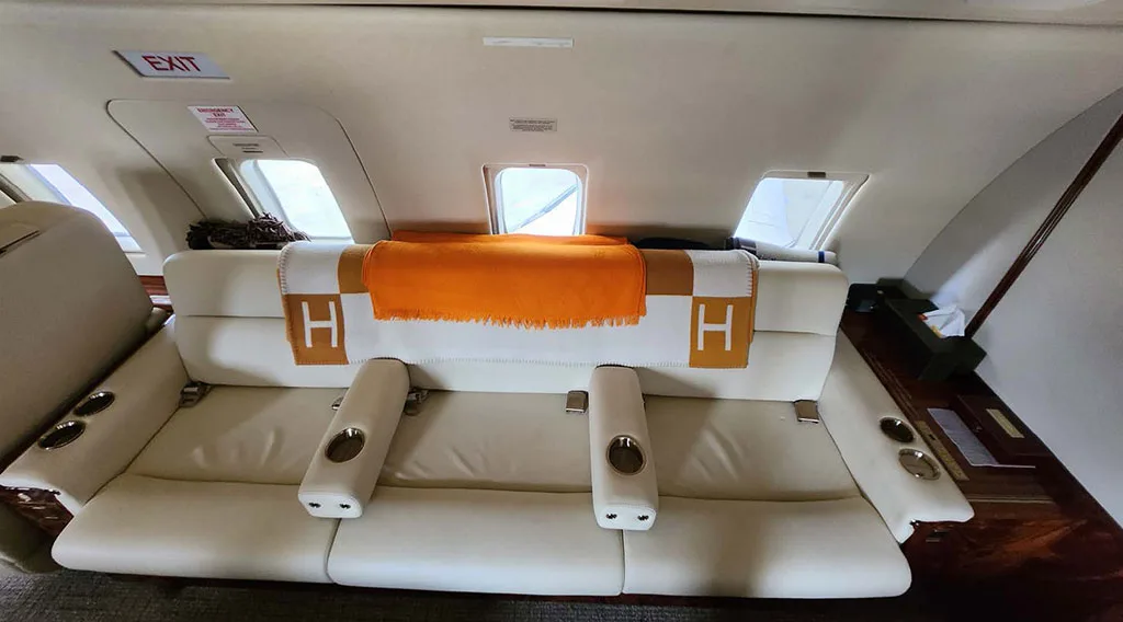 A cream leather sofa with orange and white blankets is positioned in the aircraft cabin, with an "EXIT" sign visible above.
