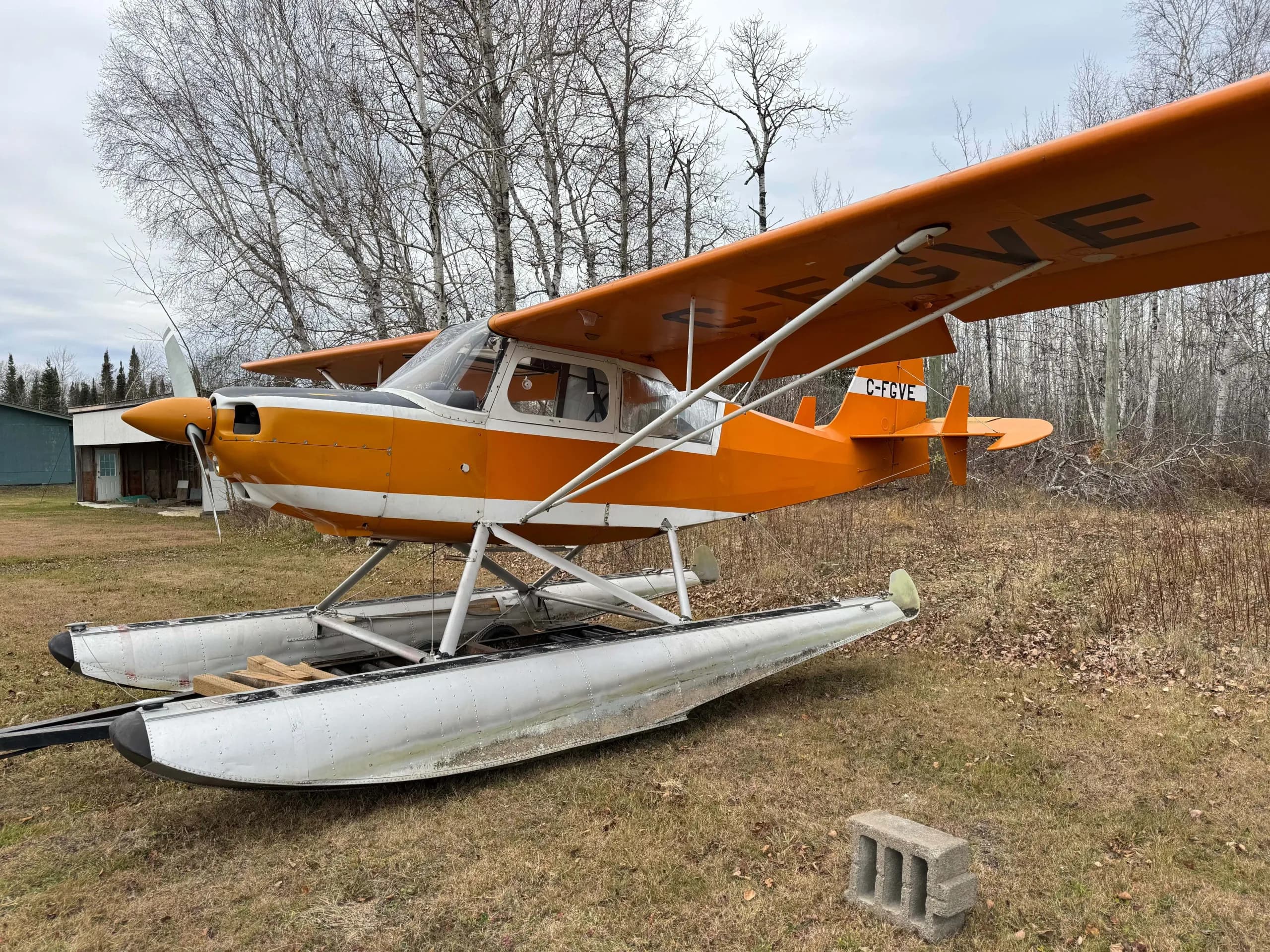 An orange and white floatplane, registration C-FGVE, parked on dry grass with bare trees and a building in the background.