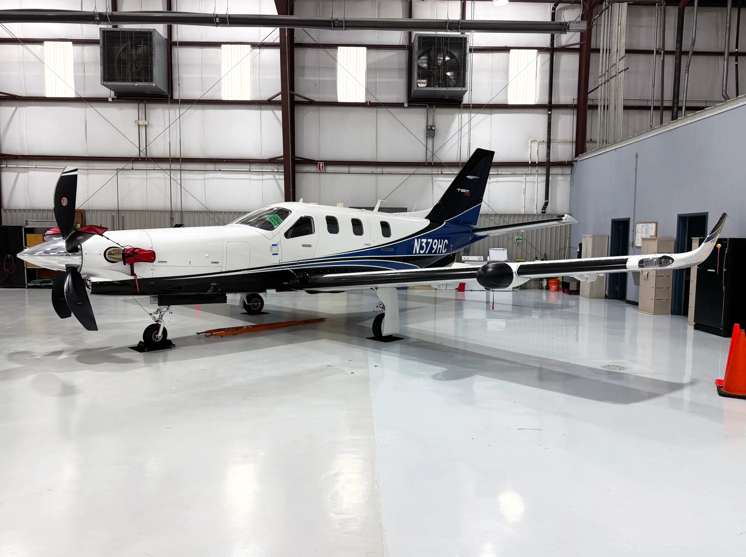 A white and blue TBM 960 aircraft with registration N379HC, parked inside a brightly lit hangar.