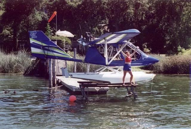 A blue and white seaplane is docked at a wooden pier with a woman standing on it, surrounded by trees and water.