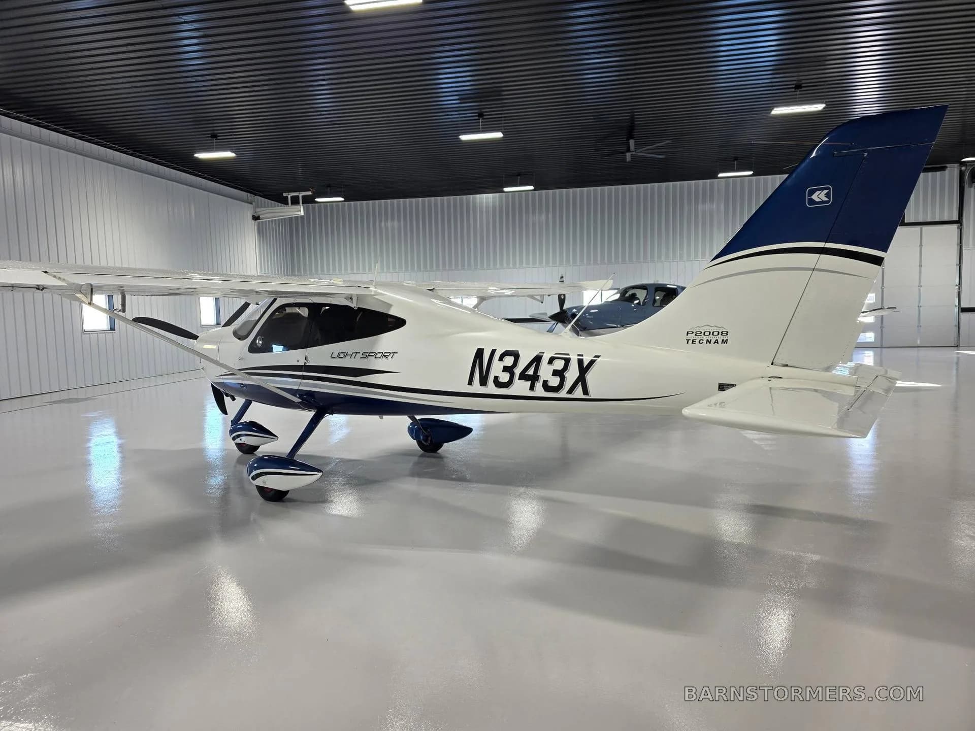 A rear-side view of a white Tecnam P2008 Light Sport aircraft with blue tail and accents, registration N343X, inside a hangar.