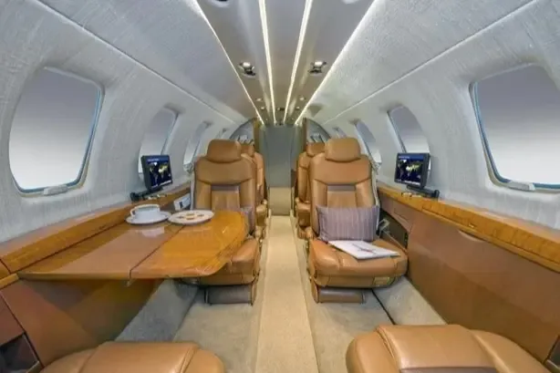 The luxurious interior of an aircraft cabin with tan leather seats, a wooden table, and overhead lighting.