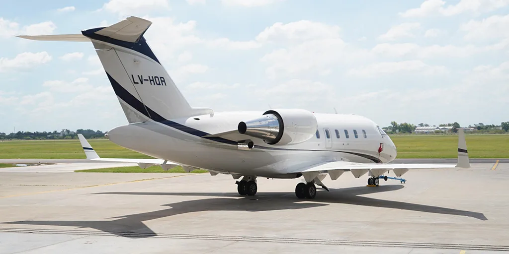 A white business jet with dark blue and grey stripes, registration LV-HQR, is parked on a tarmac with a grassy field and distant buildings under a partly cloudy sky.