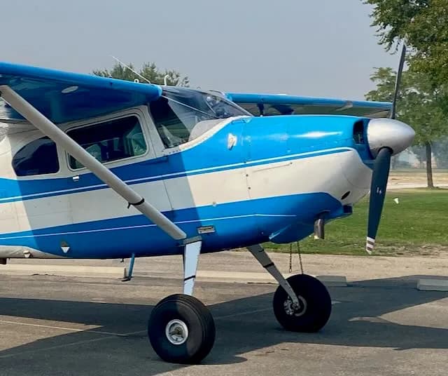 A blue and white high-wing propeller aircraft with large tires on a tarmac with trees in the background.