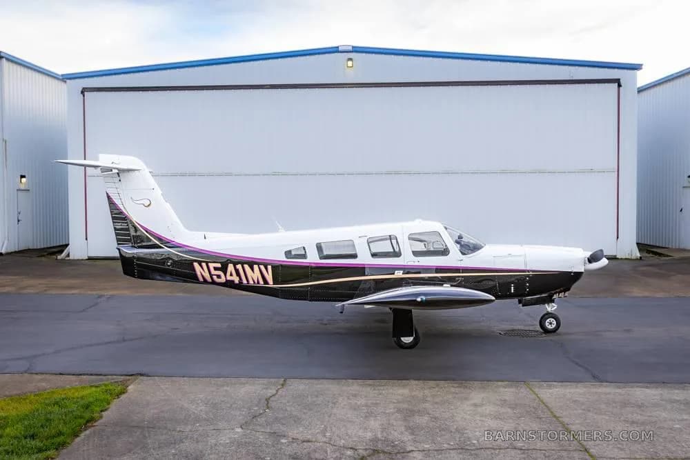 A white and black single-engine aircraft with purple stripes (N541MV) parked on an asphalt apron in front of hangars.