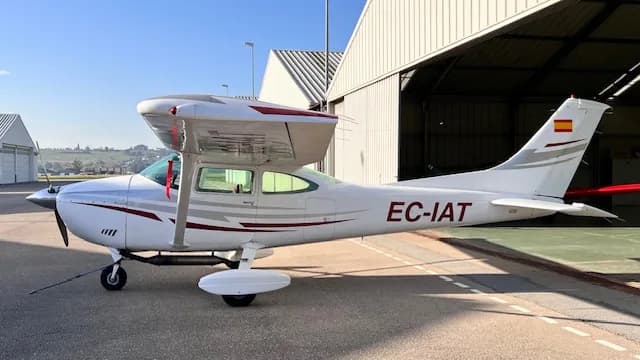 A white Cessna aircraft with red and grey stripes, registration EC-IAT, parked on an airfield in front of a hangar.