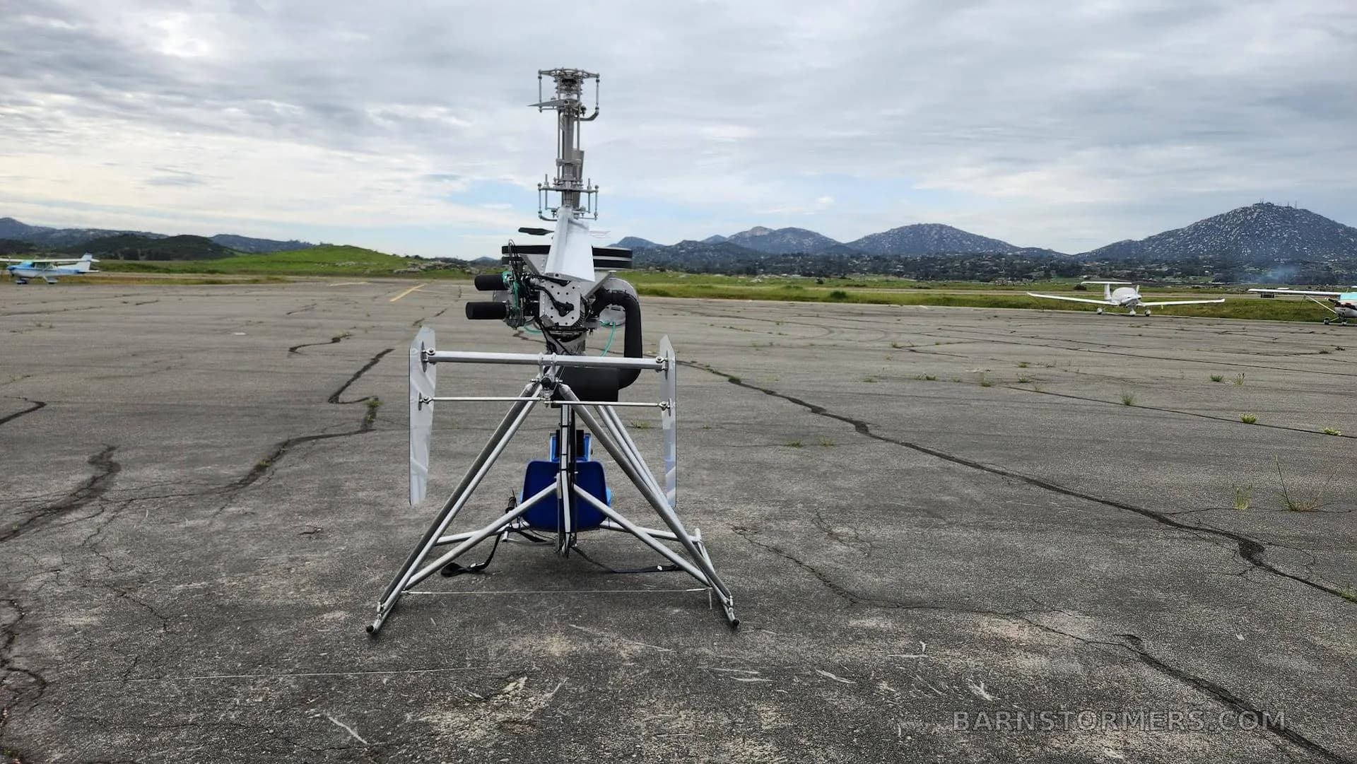A single-seat ultralight helicopter with a blue base is parked on an asphalt runway with mountains and other aircraft in the distance.