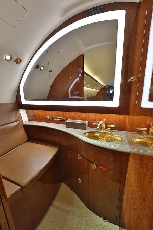 An aircraft lavatory features a gold sink, polished wood paneling, and a large mirror with accent lighting.