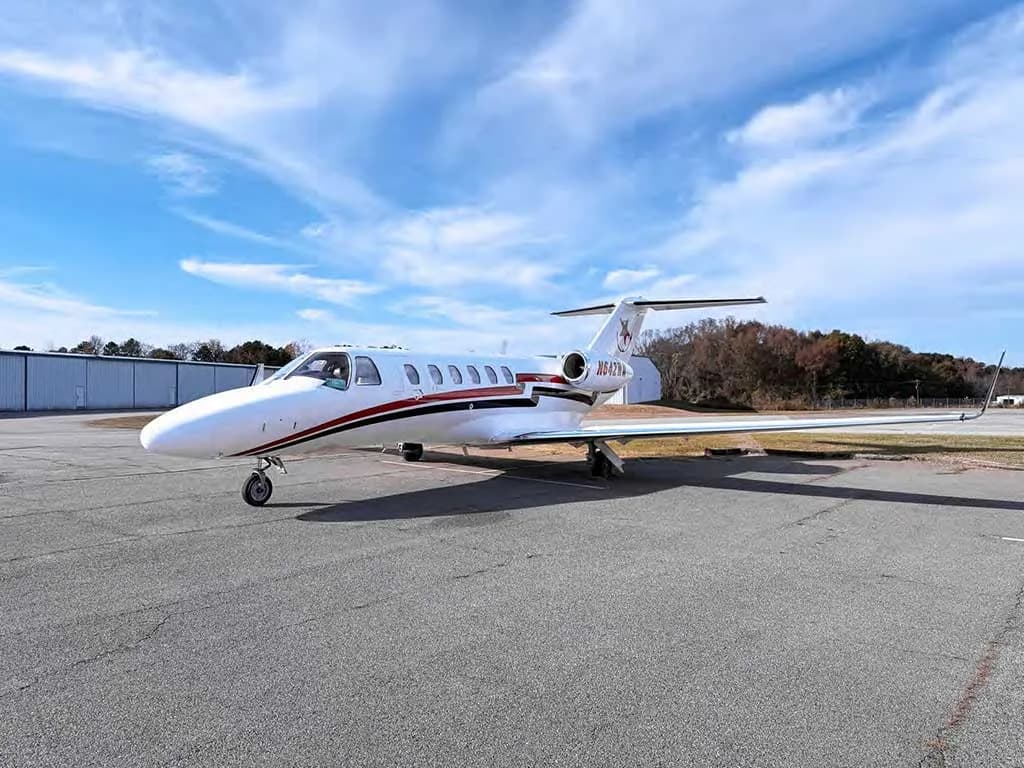 A white Cessna Citation private jet with red and black stripes and tail logo, parked on an asphalt tarmac under a partly cloudy sky.