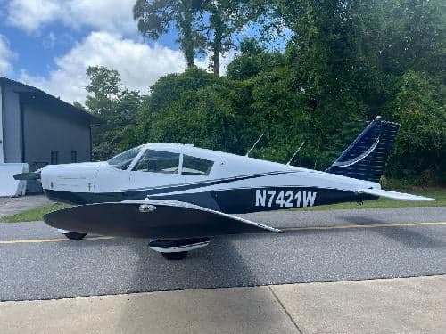 A side view of the white and dark blue Piper Cherokee 180 aircraft, registration N7421W, parked on an asphalt surface next to a building and trees.