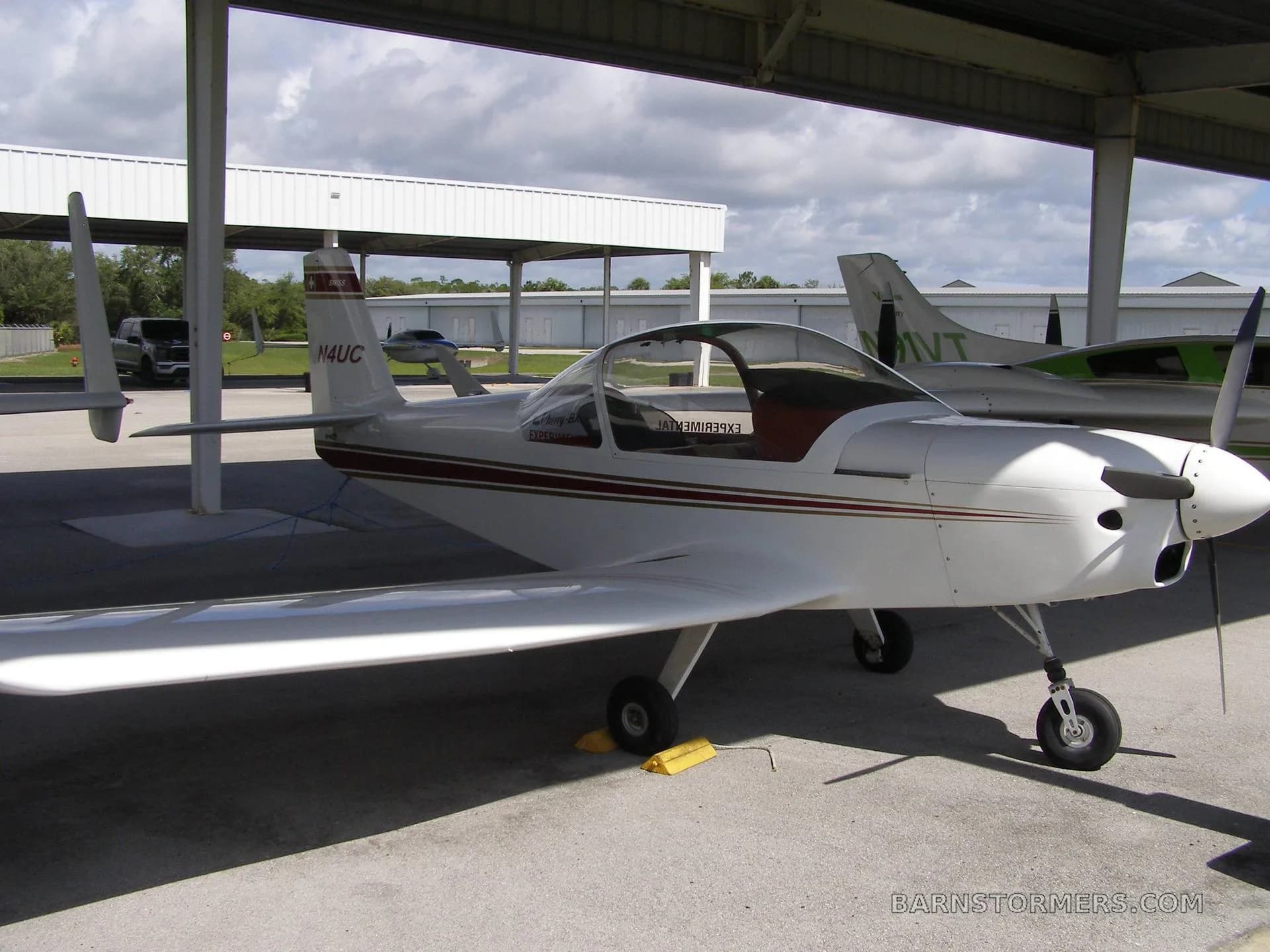 A white experimental aircraft with red and gold stripes, registration N4UC, parked under a hangar with other aircraft in the background.