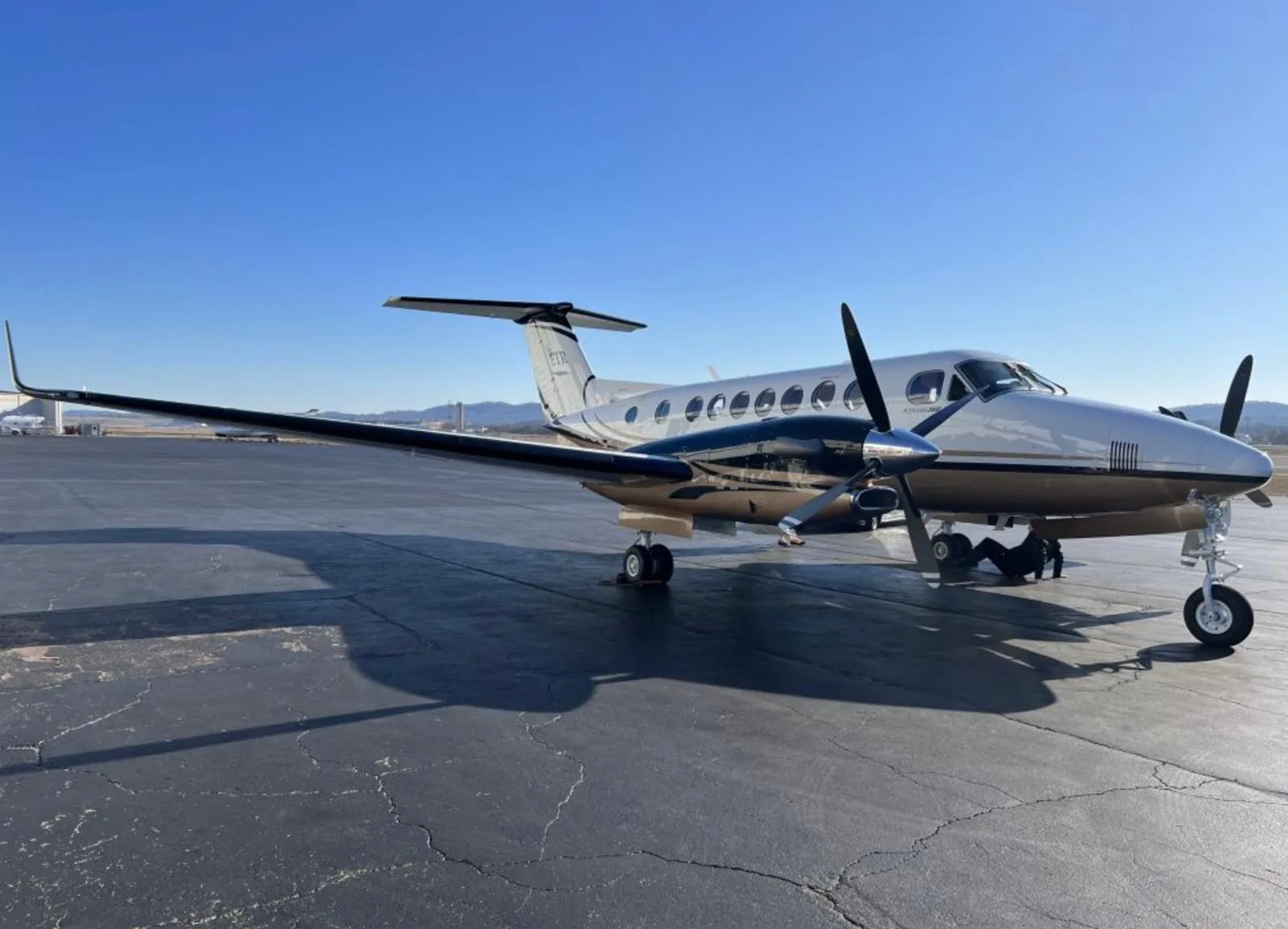 A white twin-engine turboprop aircraft with gold and blue stripes parked on a tarmac under a clear sky.