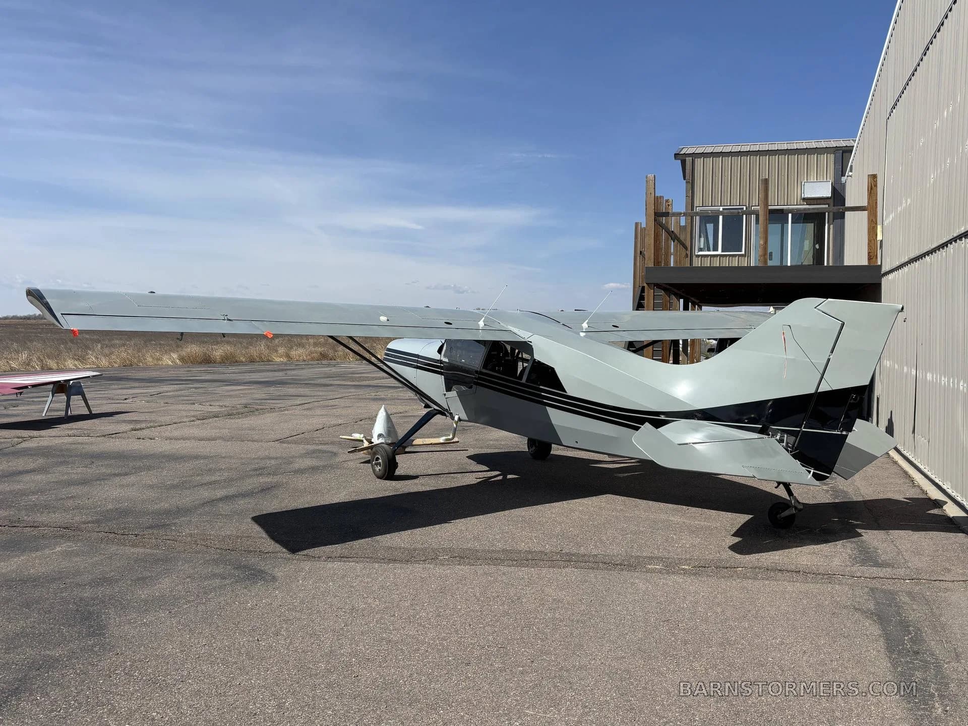 A grey Maule M-7 aircraft with black stripes parked on an asphalt ramp next to a building.